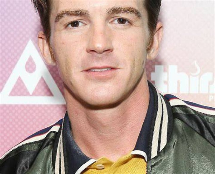 Drake Bell wearing a green puffer jacket and a yellow shirt, standing in front of a pale pink background